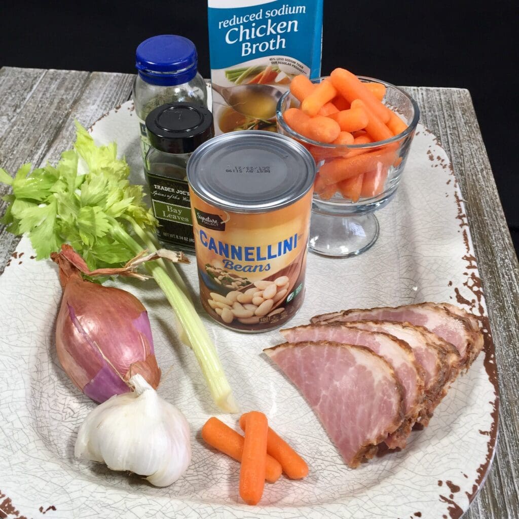 Ingredients of ham, cannellini beans, celery, carrots, garlic and chicken broth on a white platter