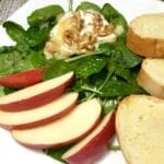 Spinach salad with sliced apples, baguette slices and toasted brie with walnuts on white plate
