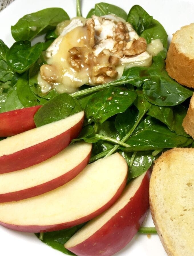 Spinach salad with sliced apples, baguette slices and toasted brie with walnuts on white plate