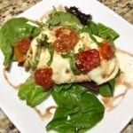 Baked Chicken with melted mozzarella and roasted tomatoes over greens on a white plate